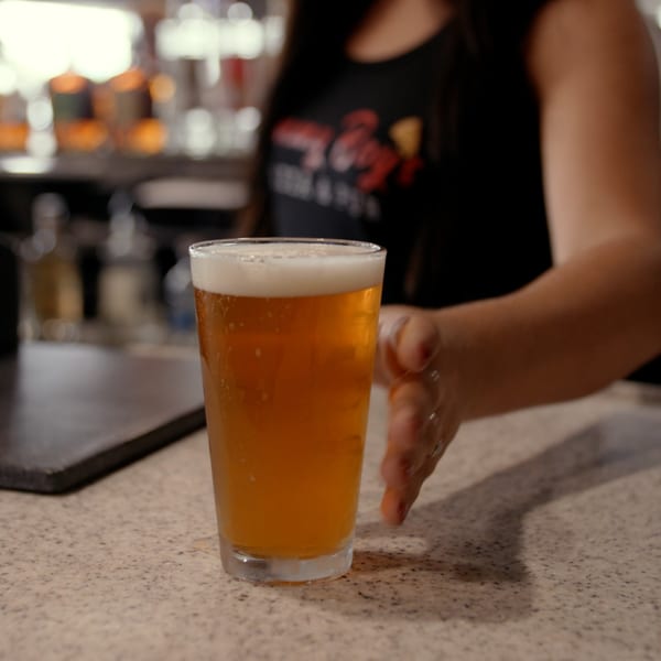 A bartender's hand slides a full glass of amber beer with a foamy head across a speckled bar.