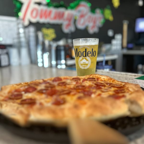 A pepperoni pizza and a Modelo beer on a counter, with a blurred "Tommy Boy's" sign in the background.