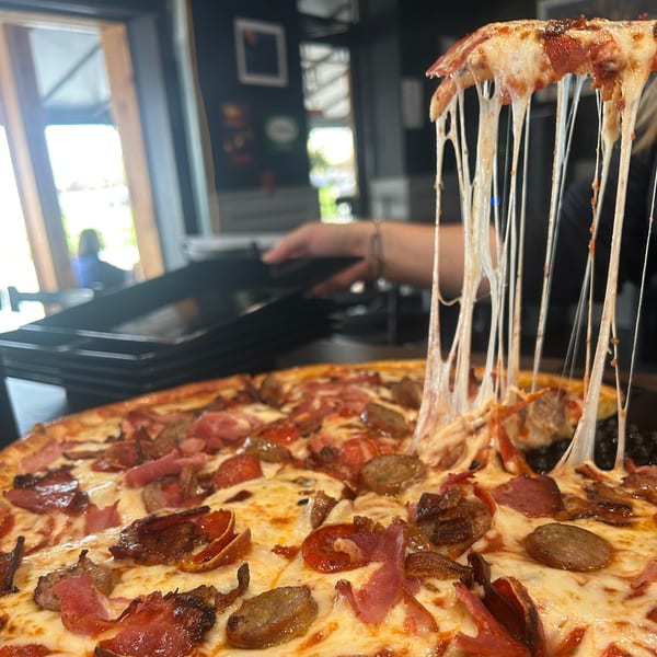 A slice of meat-lovers pizza being lifted, showing long, stretchy cheese pulls, with a restaurant interior in the background.