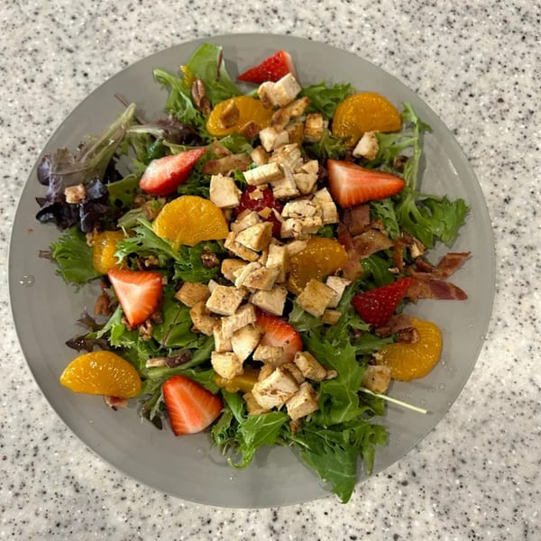 Chicken salad with mixed greens, strawberries, mandarin oranges, bacon, and pecans on a gray plate.
