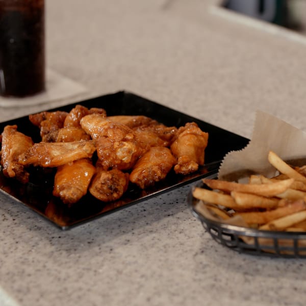A black plate of glistening chicken wings and a basket of golden french fries on a speckled countertop.