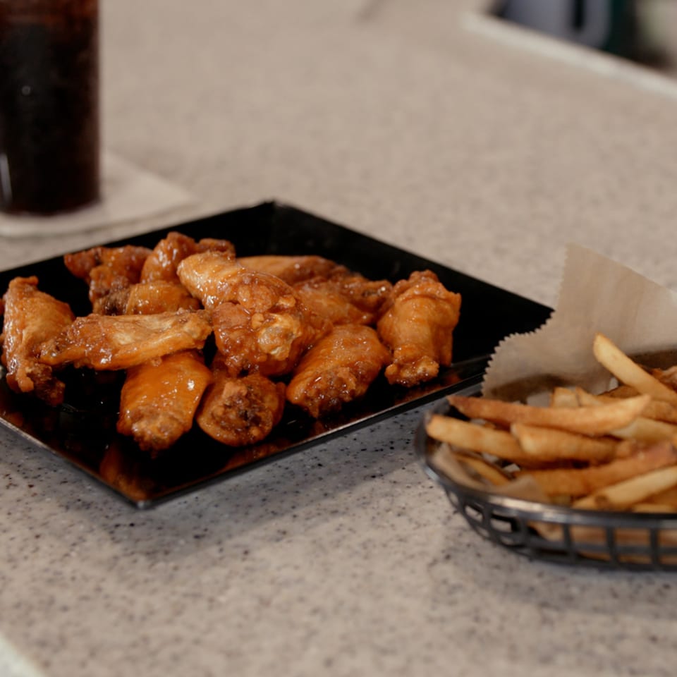 A black plate of glistening chicken wings and a basket of golden french fries on a speckled countertop.
